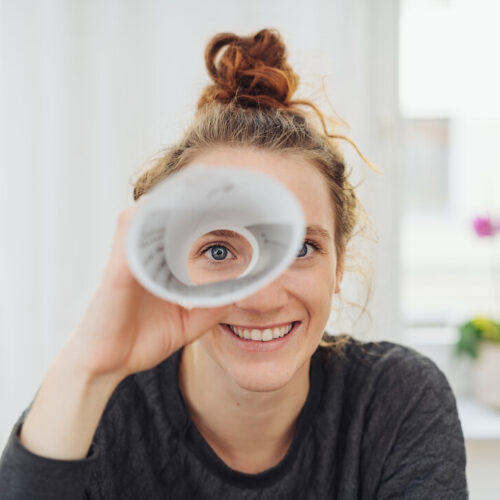 Eine Frau blickt durch ein Papierrohr in die Kamera und lächelt.