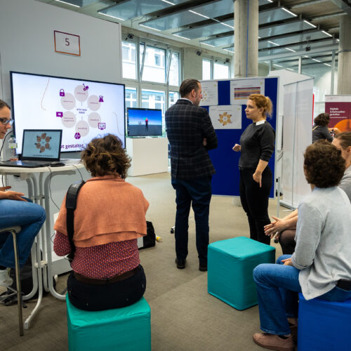 Drei Zweiergrüppchen von Menschen im Gespräch. Am Monitor im Hintergrund ist ein Bild zum Thema KI für Gute Arbeit zu sehen.