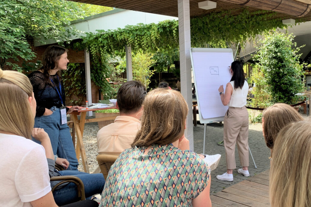 Frau präsentiert an einem Flipchart vor einer sitzenden Gruppe bei einem Workshop im Freien.