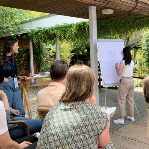 Frau präsentiert an einem Flipchart vor einer sitzenden Gruppe bei einem Workshop im Freien.
