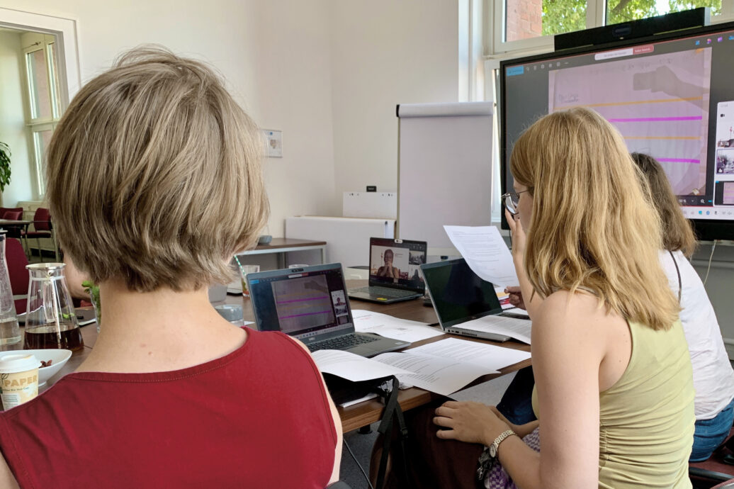 Hybride Besprechung: zwei Frauen mit Laptops vor einem Bildschirm, eine weitere Person per Videokonferenz zugeschaltet.
