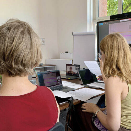 Hybride Besprechung: zwei Frauen mit Laptops vor einem Bildschirm, eine weitere Person per Videokonferenz zugeschaltet.